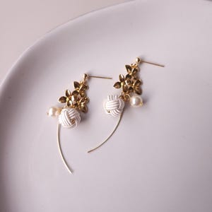 May include: A pair of gold-coloured earrings featuring floral details, white knotted elements, and faux pearls. The earrings have a delicate, curved design. The earrings are displayed on a white plate.