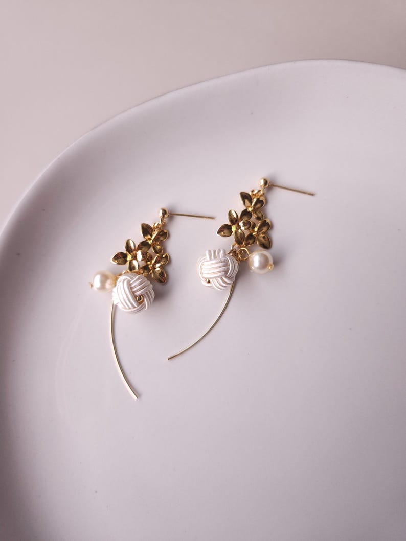 May include: A pair of gold-coloured earrings featuring floral details, white knotted elements, and faux pearls. The earrings have a delicate, curved design. The earrings are displayed on a white plate.