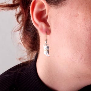 May include: A dangling earring featuring a white, knotted cord charm shaped like a cat, accented with a small gold-toned ring and a tiny white bead, suspended from a thin gold-toned metal hook that passes through the earlobe.