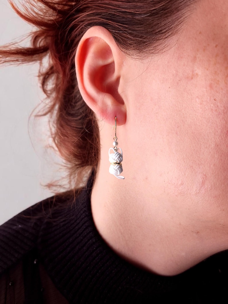 May include: A dangling earring featuring a white, knotted cord charm shaped like a cat, accented with a small gold-toned ring and a tiny white bead, suspended from a thin gold-toned metal hook that passes through the earlobe.