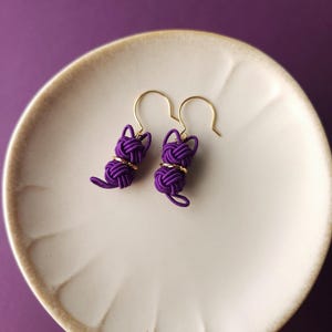 May include: A pair of purple cord earrings shaped like cats, featuring intricate knotted designs with gold-toned metal accents and gold-toned ear hooks. The earrings are crafted from thin, tightly wound purple cord, forming a head and body.
