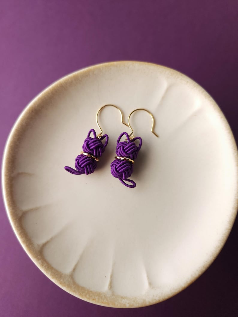 May include: A pair of purple cord earrings shaped like cats, featuring intricate knotted designs with gold-toned metal accents and gold-toned ear hooks. The earrings are crafted from thin, tightly wound purple cord, forming a head and body.