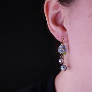 May include: Gold-tone dangle earrings featuring blue floral clusters, a gray pearl, and a patterned sphere. The earrings are designed with intricate details and a delicate aesthetic, perfect for adding a touch of elegance.