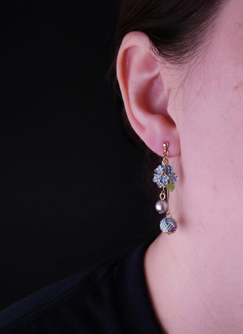May include: Gold-tone dangle earrings featuring blue floral clusters, a gray pearl, and a patterned sphere. The earrings are designed with intricate details and a delicate aesthetic, perfect for adding a touch of elegance.