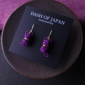 May include: A pair of purple cord earrings featuring a cat-shaped knot design with gold-tone metal accents and hooks. The earrings are attached to a black display card printed with the white text DASH OF JAPAN HANDCRAFTED.