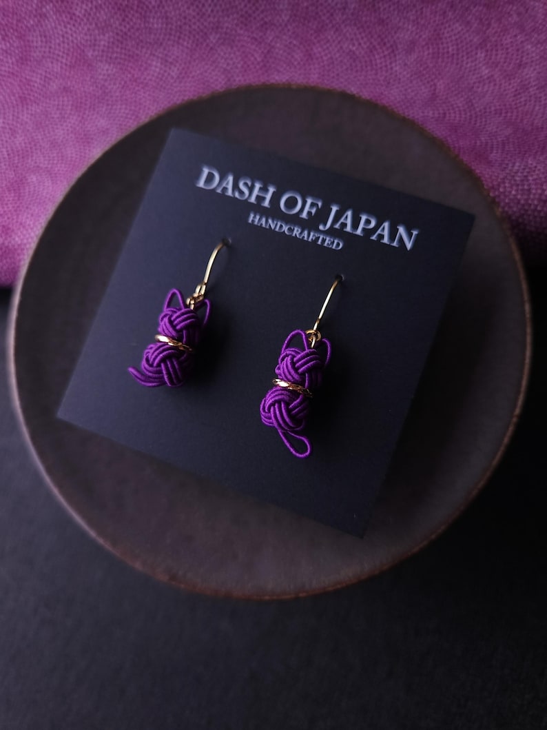 May include: A pair of purple cord earrings featuring a cat-shaped knot design with gold-tone metal accents and hooks. The earrings are attached to a black display card printed with the white text DASH OF JAPAN HANDCRAFTED.