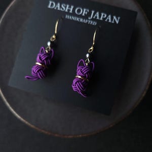 May include: A pair of purple cat-shaped earrings made from knotted cord, featuring gold-tone hooks and small dark beads. The earrings are attached to a black display card printed with the text DASH OF JAPAN HANDCRAFTED.