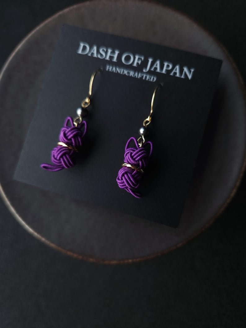 May include: A pair of purple cat-shaped earrings made from knotted cord, featuring gold-tone hooks and small dark beads. The earrings are attached to a black display card printed with the text DASH OF JAPAN HANDCRAFTED.