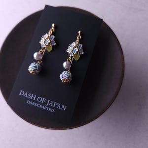 May include: A pair of handcrafted earrings featuring blue floral designs, gray pearls, and decorative knot accents. The earrings are gold-toned and displayed on a black card with the text "DASH OF JAPAN HANDCRAFTED". The earrings are on a dark brown plate.
