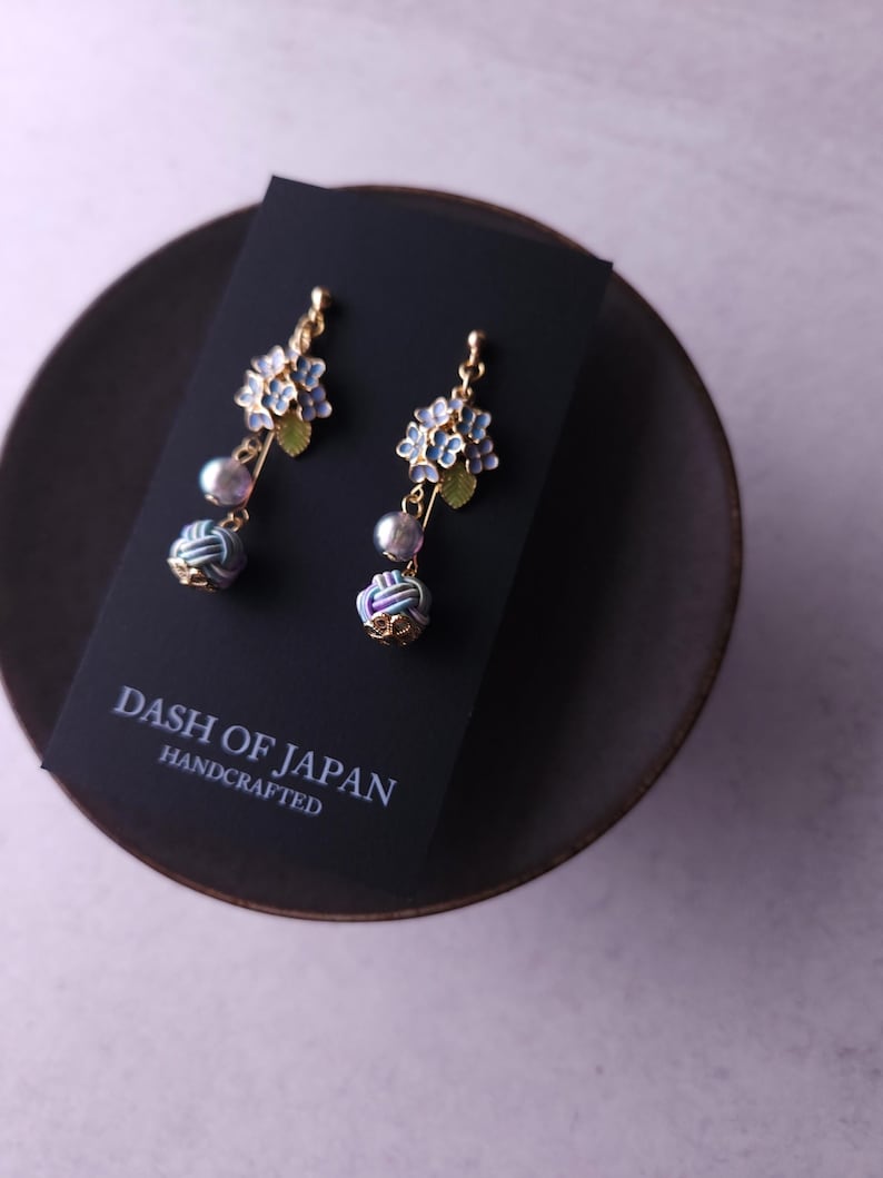 May include: A pair of handcrafted earrings featuring blue floral designs, gray pearls, and decorative knot accents. The earrings are gold-toned and displayed on a black card with the text "DASH OF JAPAN HANDCRAFTED". The earrings are on a dark brown plate.