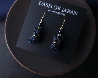 May include: A pair of dark blue cat-shaped earrings made from woven cord, featuring gold-tone metal accents and small dark blue beads. The earrings are attached to a black display card with the white text DASH OF JAPAN HANDCRAFTED printed on it.