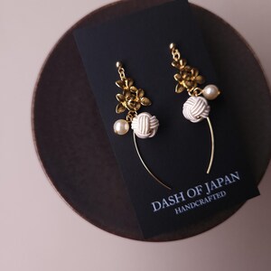 May include: Gold-coloured floral earrings with white knotted details and faux pearls. The earrings are presented on a black card with the text "DASH OF JAPAN HANDCRAFTED". The earrings are on a dark brown surface.