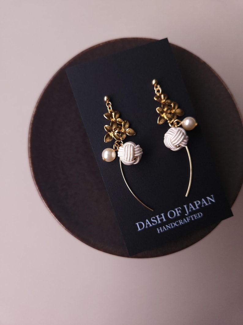 May include: Gold-coloured floral earrings with white knotted details and faux pearls. The earrings are presented on a black card with the text "DASH OF JAPAN HANDCRAFTED". The earrings are on a dark brown surface.