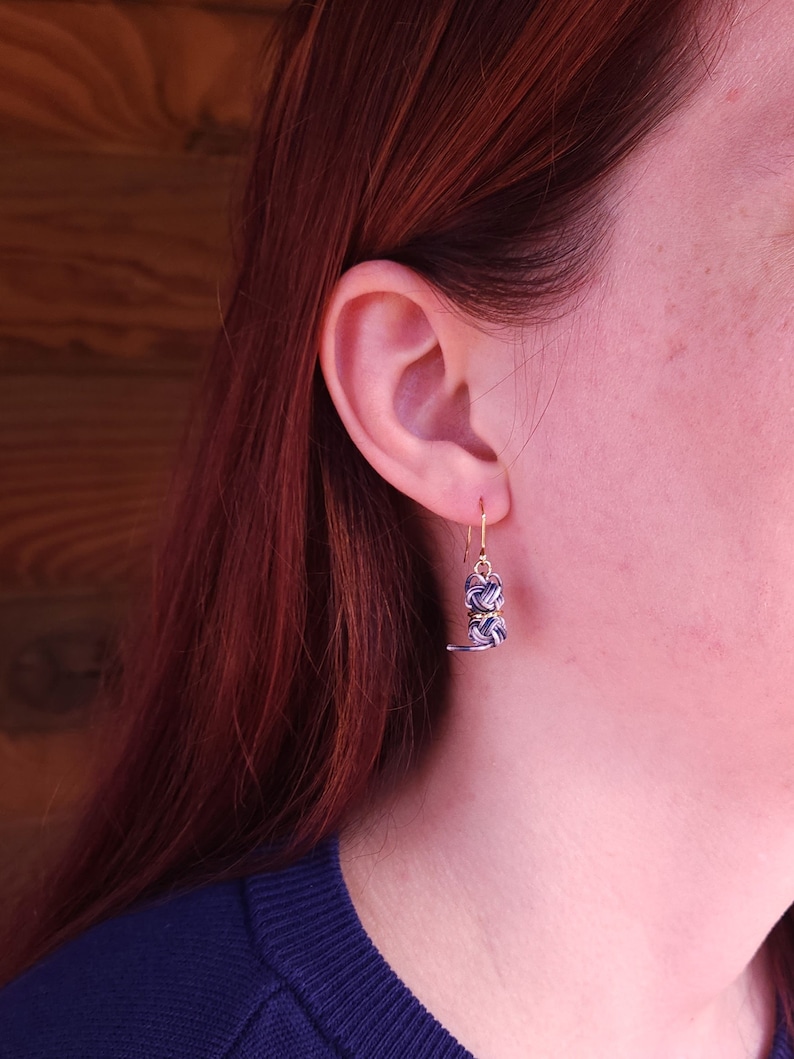 May include: A dangling earring featuring a small, spherical knot design crafted from thin, intertwined navy blue and white cord, accented with metallic gold wire, suspended from a simple gold-toned metal hook wire.