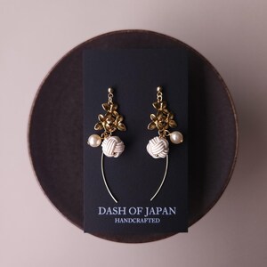 May include: A pair of gold-coloured floral earrings with white knotted details and faux pearls. The earrings are presented on a black card with the text "DASH OF JAPAN HANDCRAFTED". The earrings hang from a curved gold wire.