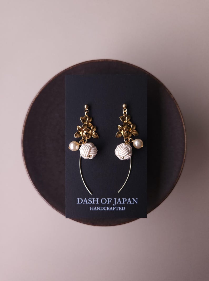 May include: A pair of gold-coloured floral earrings with white knotted details and faux pearls. The earrings are presented on a black card with the text "DASH OF JAPAN HANDCRAFTED". The earrings hang from a curved gold wire.