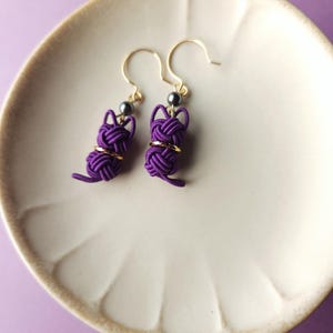 May include: A pair of purple cat-shaped earrings crafted from knotted cord. Each earring features a small dark bead at the top, a gold-toned ring around the middle, and a dangling tail. The earrings hang from gold-toned metal hooks.