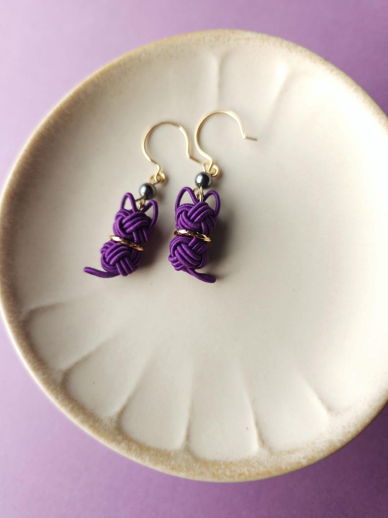 May include: A pair of purple cat-shaped earrings crafted from knotted cord. Each earring features a small dark bead at the top, a gold-toned ring around the middle, and a dangling tail. The earrings hang from gold-toned metal hooks.