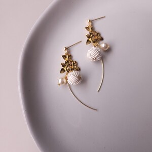 May include: A pair of elegant earrings with gold-coloured floral accents, white knotted details, and small pearl-like beads. The earrings have a delicate, curved design, with a post-back closure.
