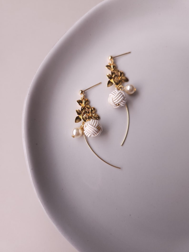 May include: A pair of elegant earrings with gold-coloured floral accents, white knotted details, and small pearl-like beads. The earrings have a delicate, curved design, with a post-back closure.