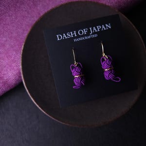 May include: A pair of purple cord knot earrings with gold-tone metal hooks, displayed on a black card labeled DASH OF JAPAN HANDCRAFTED. The earrings feature intricate, woven purple cord designs shaped into small, rounded, knotted forms.