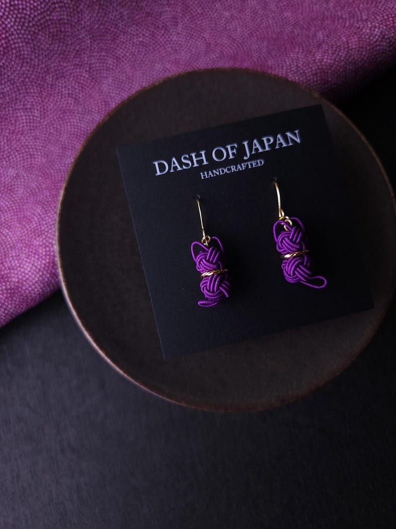 May include: A pair of purple cord knot earrings with gold-tone metal hooks, displayed on a black card labeled DASH OF JAPAN HANDCRAFTED. The earrings feature intricate, woven purple cord designs shaped into small, rounded, knotted forms.