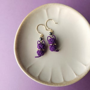 May include: A pair of purple cord earrings featuring a knotted cat-shaped design. Each earring includes a small metallic bead and a gold-toned hook wire. The earrings are crafted from thin, tightly wound purple cord, forming a distinct feline silhouette.