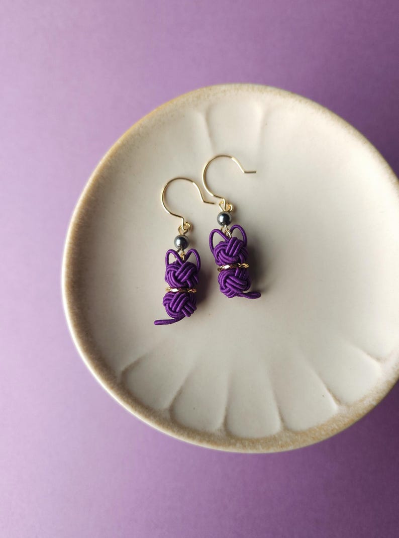 May include: A pair of purple cord earrings featuring a knotted cat-shaped design. Each earring includes a small metallic bead and a gold-toned hook wire. The earrings are crafted from thin, tightly wound purple cord, forming a distinct feline silhouette.