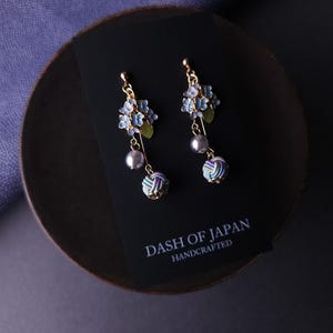 May include: Gold-toned earrings featuring blue floral clusters, green leaves, and dangling gray pearls. Below the pearls are decorative, multi-colored knot-like accents. The earrings rest on a black card with the text "DASH OF JAPAN HANDCRAFTED".