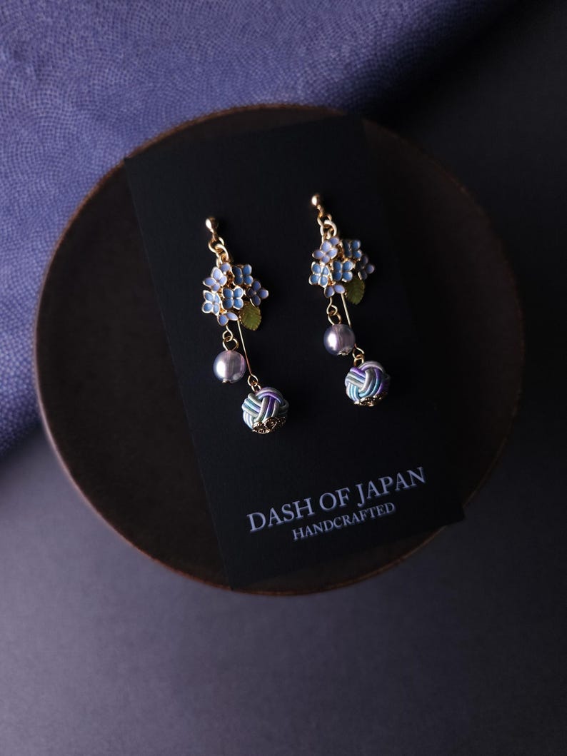May include: Gold-toned earrings featuring blue floral clusters, green leaves, and dangling gray pearls. Below the pearls are decorative, multi-colored knot-like accents. The earrings rest on a black card with the text "DASH OF JAPAN HANDCRAFTED".
