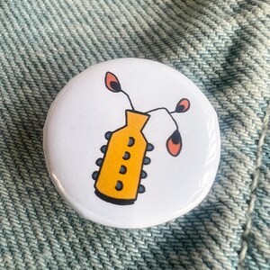 May include: A circular pinback button featuring a white background with a central illustration of a yellow vase with blue circular accents along its sides. Two thin black stems emerge from the top, each ending in a small, rounded orange leaf shape.
