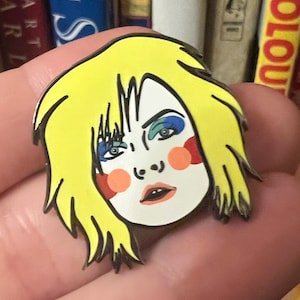 Op de afbeelding: Een gele emaille pin met een cartoonportret van een persoon met blond haar, blauwe en groene ogen en rode en oranje blos op de wangen.