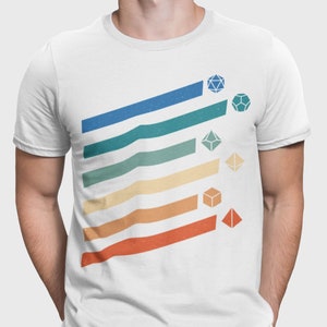 Puede incluir: Una camiseta blanca con un diseño de estilo retro con rayas de colores y dados. Los dados son de diferentes colores y formas, y están dispuestos en una línea diagonal.