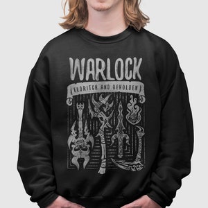 Op de afbeelding: Zwart sweatshirt met een witte grafische print van toverspullen, waaronder een schedel, een staf en een vlam. De tekst "Warlock" en "Eldritch and Beholder" staat op de print.