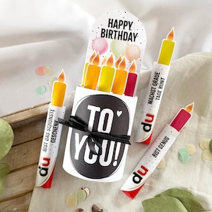 May include: A birthday-themed gift set featuring a box with "TO YOU!" text, a "HAPPY BIRTHDAY" card, and candle-shaped items. The candles are white with colorful tops and flames. The set includes confetti and a black ribbon.