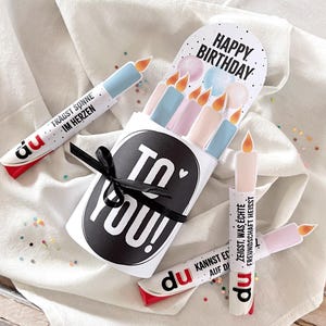 May include: A birthday-themed gift set featuring candle-shaped paper tubes with text and a black ribbon. The set includes a card that says "Happy Birthday" and candles in pastel colors. The tubes have German text.