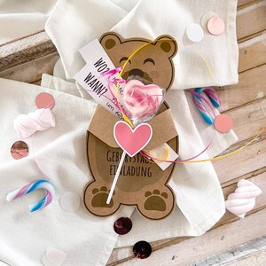 May include: A brown teddy bear-shaped invitation card with the text "Geburtstag Einladung" and a pink heart. A heart-shaped lollipop is attached. The card is on a white cloth with confetti and candies.