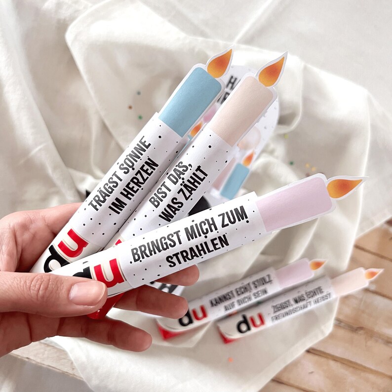 May include: Several white paper candle-shaped decorations with colorful tops and flame cutouts. Each candle has a different message in German, such as "Brings me to shine." The brand name "du" is printed on the bottom of each candle.