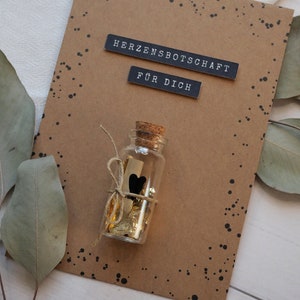 May include: A brown card with black polka dots and the text "Herzensbotschaft f&uuml;r dich" in black lettering. A small glass bottle with a cork top is tied with twine and filled with gold foil and a small black heart. The bottle is sitting on the card.