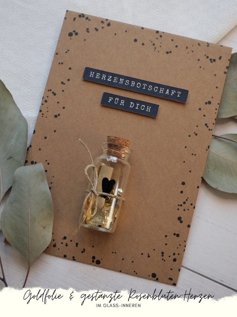 May include: A brown card with black polka dots and the text "Herzensbotschaft f&uuml;r dich" in black lettering. A small glass bottle with a cork top is tied with twine and filled with gold foil and a small black heart. The bottle is sitting on the card.