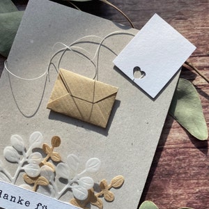 May include: A handmade greeting card with a small brown paper envelope, a white card with a heart cutout, and decorative floral elements. The card is on a gray background with a string and leaves.