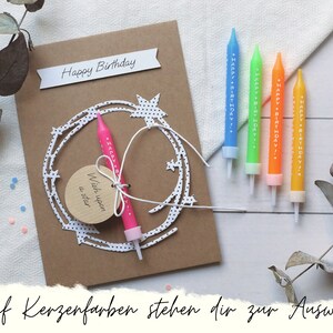 May include: A brown card with the text "Happy Birthday" and a white paper cutout of a star wreath. A pink birthday candle with the text "Happy Birthday" is attached to the card. The card is on a white surface with five birthday candles in different colors.