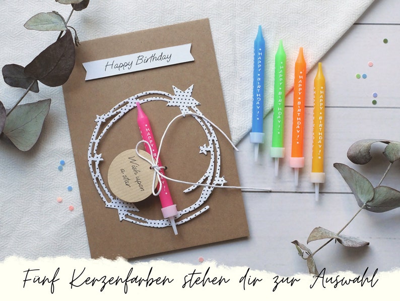May include: A brown card with the text "Happy Birthday" and a white paper cutout of a star wreath. A pink birthday candle with the text "Happy Birthday" is attached to the card. The card is on a white surface with five birthday candles in different colors.