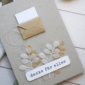 May include: A handmade greeting card with a kraft paper envelope and a white card peeking out. The card features white and tan paper leaf cutouts and a white label that reads "danke f&uuml;r alles".