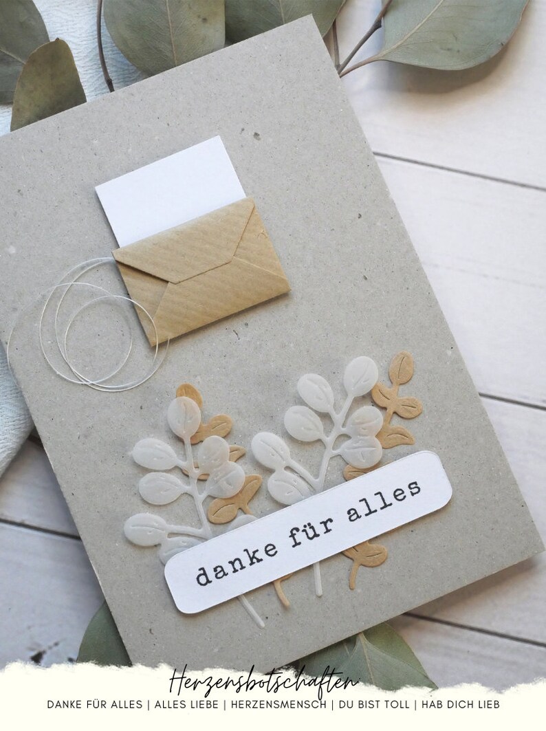 May include: A handmade greeting card with a kraft paper envelope and a white card peeking out. The card features white and tan paper leaf cutouts and a white label that reads "danke f&uuml;r alles".