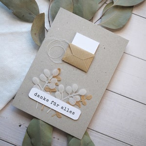 May include: A handmade greeting card with a kraft paper envelope and a white card peeking out. The card is gray with white and tan leaf embellishments and the text "danke f&uuml;r alles". Eucalyptus leaves are arranged around the card.