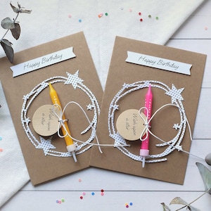 May include: Two brown cardstock birthday cards with white polka dot stars and a white string with a small wooden tag that says "Wish upon a star". Each card has a birthday candle, one yellow and one pink, attached to the string.