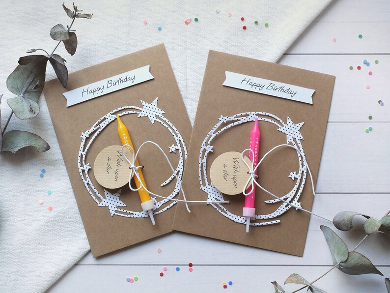 May include: Two brown cardstock birthday cards with white polka dot stars and a white string with a small wooden tag that says "Wish upon a star". Each card has a birthday candle, one yellow and one pink, attached to the string.