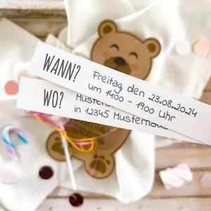 May include: A light brown teddy bear-shaped tag with the text "WANN?" and "WO?" is paired with two white strips of paper with handwritten text. The text includes the date, time, and location. A lollipop and confetti are also visible.