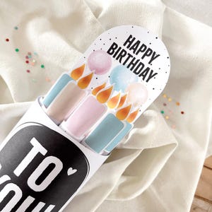 May include: A white paper tube with the words "TO YOU" in black. Inside are pastel-colored candle-shaped cards with the words "HAPPY BIRTHDAY" on top. The candles have orange flames and are pink, blue, and beige.